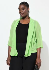 Giacca aperta trasparente verde lime con maniche a tre quarti, indossata sopra un top nero e abbinata a pantaloni neri con tessuto testurizzato.