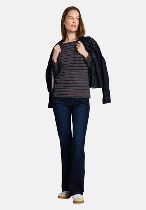 Vrouw met een gestreept shirt met lange mouwen, donkere jeans en witte sneakers, die een donkere jas over haar schouders draagt, staand tegen een witte achtergrond.