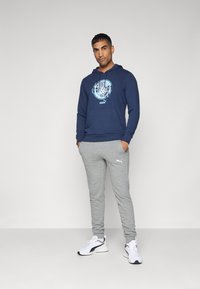 Felpa navy con stampa grafica, abbinata a pantaloni sportivi grigi chiari e scarpe da ginnastica bianche. Il cappuccio è dotato di lacci regolabili.