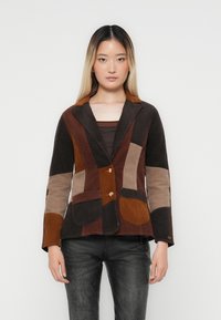 Bruine corduroy patchwork blazer met contrasterende zakken, voorzien van een geknopt kraag en twee knopen. G gedragen over een donkere top.