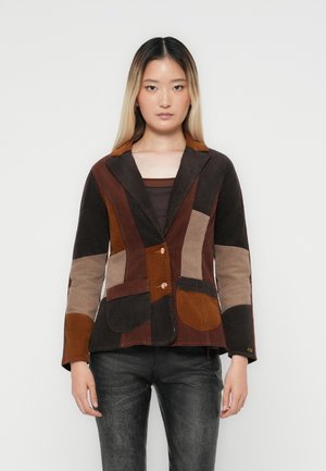 Brązowa marynarka z kordurowego materiału w stylu patchwork, z kontrastowymi kieszeniami, z klapami i dwoma guzikami. Nosi ją na ciemnej bluzce.
