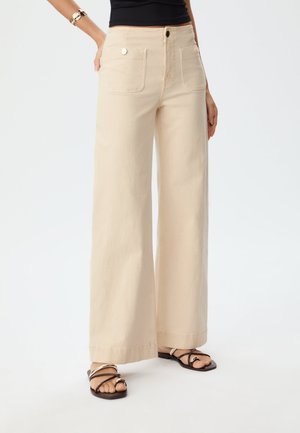 Pantalon large beige en coton, doté de deux poches avant et d'une fermeture à bouton. L'ourlet est proprement fini. Porté avec des sandales noires.