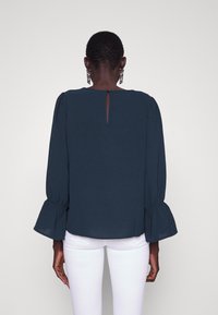 Vrouw met haar rug naar de camera gekeerd, gekleed in een donkerblauwe blouse met lange mouwen en uitlopende mouwen, en witte strakke broeken, tegen een effen lichte achtergrond.