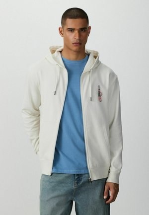 Junger Mann, der einen weißen Zip-up-Hoodie mit einem Flaschenaufnäher über einem blauen T-Shirt und hellblauen Jeans trägt, steht vor einem schlichten weißen Hintergrund.