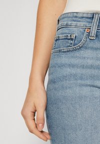 Vaaleansiniset farkut, joissa on vartalonmyötäinen leikkaus. Yksityiskohtiin kuuluu etutasku, ompelut ja kuparinvärinen nappi vyötäröllä.