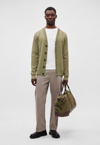 Olivovo zelený rebrovaný kardigán s čiernymi gombíkmi, biele tričko, svetlo béžové nohavice a čierne topánky. Drží zeleno-hnedú športovú tašku.