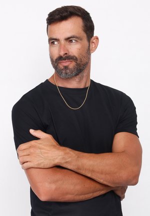 Black fitted t-shirt, short sleeves, crew neck, worn by a man with a gold chain around neck, arms crossed against a plain white background.