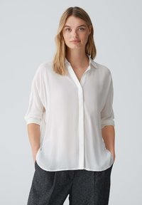 Blusa bianca e leggera con bottoni frontali, maniche arrotolate e orlo curvo, abbinata a pantaloni scuri e strutturati.