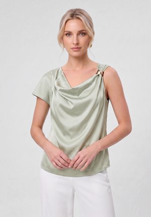 Vrouw met blond haar die een lichtgroene satijnen asymmetrische top met gouden gesp en witte broek draagt, staand tegen een effen achtergrond.