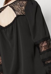 Svart blus med blommiga spetsdetaljer längs ärmarna och ryggen. Mjuk tyg med en lätt glans och en rundad halsringning.