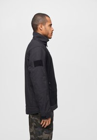 Black jacket with high collar, textured fabric, side zip pockets, and a Velcro patch on left sleeve. Worn over camo cargo pants.
