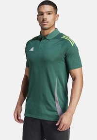 Polo verde con maniche corte, realizzato in tessuto leggero. Presenta tre righe gialle sulla spalla e un pannello laterale grigio.