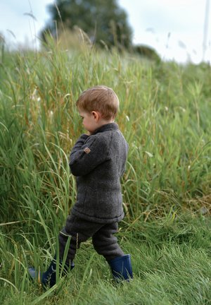 Child wearing a dark grey woolen outfit with rolled cuffs and blue rubber boots, standing in tall green grass. Back view.
