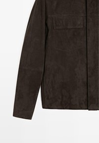 Giacca in suede marrone dalla texture liscia, maniche lunghe e taschino sul petto. Presenta un design minimalista con linee pulite e senza elementi hardware visibili.