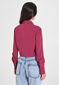 Femme aux longs cheveux noirs portant un blouse à manches longues framboise rentrée dans un jean taille haute bleu clair, se tenant devant un fond neutre.