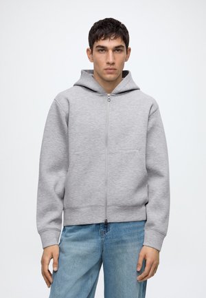 Jeune homme faisant face, portant un sweat à capuche zippé gris clair et un jean bleu sur un fond blanc uni.