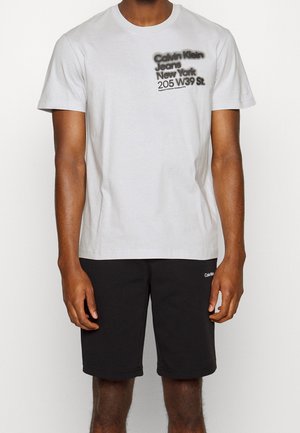 Man draagt een wit Calvin Klein T-shirt met tekst op de linker borst en zwarte korte broek met klein logo op linker dij.