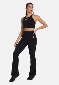 Black sports bra with cut-out detail, paired with high-waisted black flared leggings. Both made from smooth, stretchy fabric; logo accents visible.