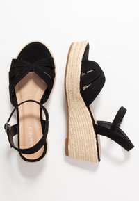 Sandales compensées en suède noir avec des brides entrecroisées et une boucle à la cheville. Semelle en espadrille en fibre naturelle, semelle intérieure en cuir tan. Talon texturé.