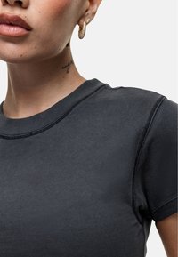T-shirt aderente nera con scollatura rotonda, maniche corte e dettagli di cucitura visibili. Il modello mostra un tatuaggio sul collo e orecchini a cerchio.