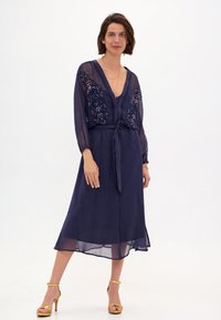 Robe bleu marine avec des manches longues transparentes, des broderies florales sur le corsage, un décolleté en V et une taille nouée. Tissu lisse, longueur cheville.
