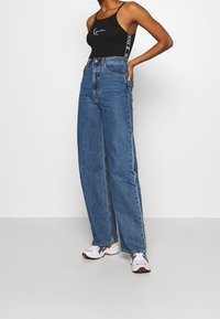 Top senza maniche nero con logo bianco, abbinato a jeans blu in denim a vita alta e gamba larga, e scarpe sportive bianche con dettagli neri.