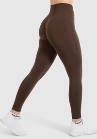 Bruine hoge taille legging met een gladde textuur en taps toelopende pasvorm, gecombineerd met witte sportschoenen met een zwart logo op de sok.