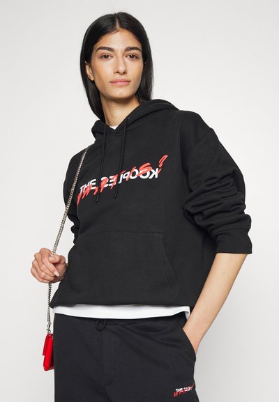 Schwarzer Hoodie mit einer Fronttasche, versehen mit fettem weißen und roten Text. Kombiniert mit schwarzen Hosen und einem kleinen roten Accessoire. Lässiger Schnitt.