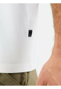 Hvid bomulds T-shirt med et sort mærke på venstre side. T-shirten har en glat tekstur og en afslappet pasform, som er parret med olivenfarvede bukser.