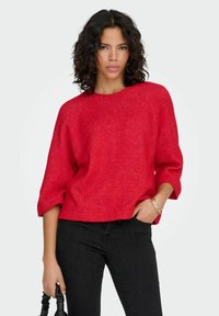 Rdeč pulover s teksturirano pletenino, z ladjskim izrezom in rokavi do komolcev, v kombinaciji s črnimi kavbojkami. Preprosta, vsakdanja zasnova.