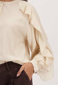 Blouse beige à manches longues avec des détails en volants le long des manches et des poignets, assortie à un pantalon marron foncé, main dans la poche.