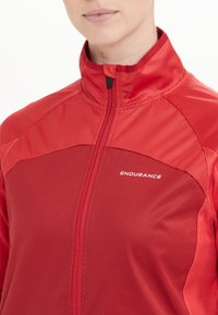 Röd sportjacka med hög krage, dragkedjestängning och textil med struktur. Har "ENDURANCE"-logotypen i vitt på bröstet.