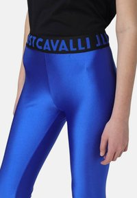 Blauwe leggings van een glanzend, soepel materiaal. Ze hebben een zwarte tailleband met "JUST CAVALLI" in het blauw gedrukt.