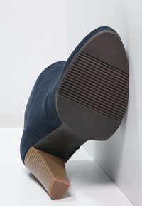 Chaussure à talon haut en daim bleu marine avec une semelle en caoutchouc texturé et un talon rectangulaire beige, arborant de subtils détails de couture.