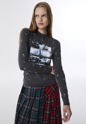 Grijze lange mouw shirt met parelversieringen en een filmstrip grafiek. Gecombineerd met een veelkleurige geruite rok met groene en rode segmenten.