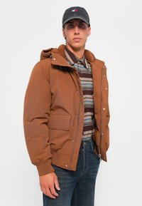 Brązowa ocieplana kurtka z kapturem, z przednim zamkiem błyskawicznym, napami i zewnętrzną kieszenią. Nosi na sobie paskowaną koszulę zapinaną na guziki.