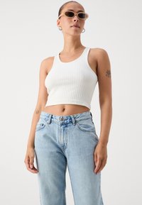 Λευκή ριμπ μπλούζα κοντή με φαρδιές τιράντες, συνδυασμένη με ανοιχτό μπλε τζιν ψηλόμεσα. Το μοντέλο φορά μινιμαλιστικά γυαλιά ηλίου και σκουλαρίκια.