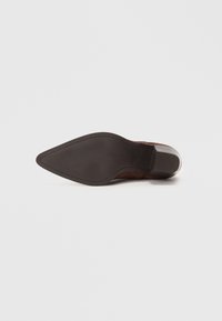 Botín de ante marrón con tacón, punta afilada, que presenta una suela negra lisa y un tacón cuadrado. Detalles mínimos, diseño limpio.