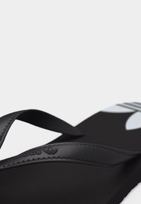 Κοντινό πλάνο μαύρης σαγιονάρας Adidas με υφασμένη λουρίδα και λευκό λογότυπο στο πέλμα πάνω σε απλό φόντο.