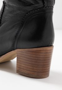 Bottine en cuir noir avec un talon en bois superposé. Présente des détails de couture visibles et une texture lisse sur le dessus.