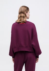 Persona con capelli ondulati alla spalle che indossa una felpa viola larga e pantaloni abbinati, vista di spalle su uno sfondo bianco.