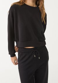 Schwarzes Sweatshirt mit überschnittenen Schultern und verkürztem Saum, hergestellt aus weichem Stoff. Kombiniert mit passenden schwarzen Jogginghosen mit elastischem Bund.