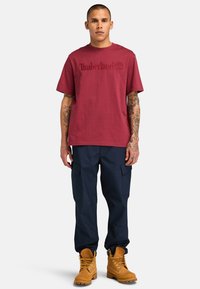 Camiseta de algodón burdeos con el logotipo en relieve de "Timberland", combinada con pantalones cargo azul marino y botas impermeables color miel con cordones amarillos.