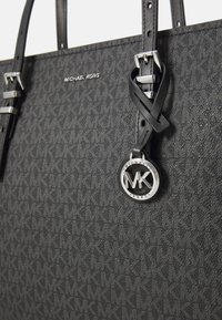 Melns Michael Kors somiņa ar teksturētu dizainu, sudraba aksesuāriem un apļveida logotipa birku, kas piestiprināta pie roktura.