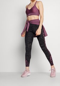 Sport-BH in Pflaume mit dünnen Trägern, kombiniert mit hoch taillierten schwarzen Leggings mit einem strukturierten floralen Muster und pinken Sportschuhen.