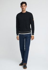 Svart kabelstickad tröja med rund halsringning, vit kant vid midjan och ärmslut, tillsammans med mörk jeans och bruna skor.