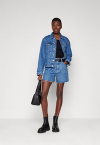 Chaqueta de mezclilla y pantalones cortos en azul, combinados con una camiseta sin mangas negra y cinturón. Botines negros y un pequeño bolso negro completan el look.