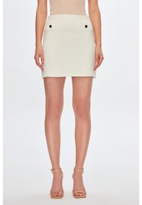 Witte textuur mini rok met twee voorzakken en zwarte knopen. De stof heeft een subtiel geweven patroon, wat zorgt voor een gestructureerde uitstraling.