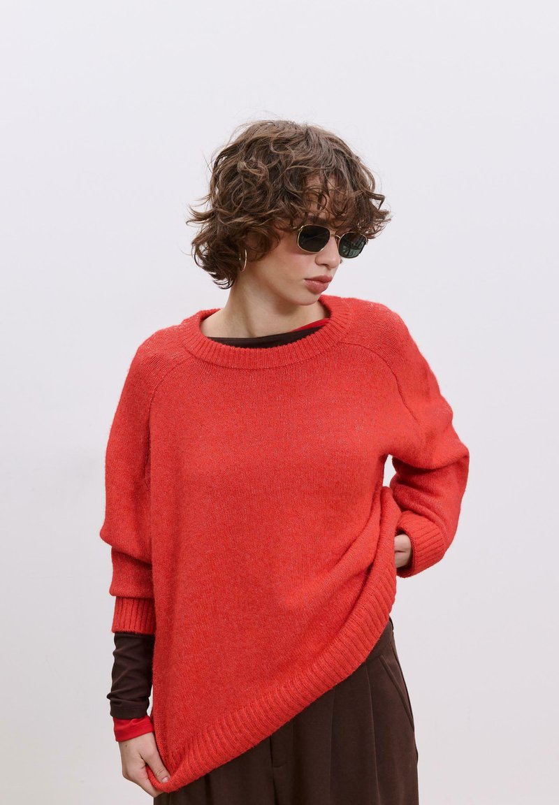 Pull en maille rouge à coupe décontractée, col large et ourlet asymétrique. Associé à un haut à manches longues foncé et des lunettes de soleil.