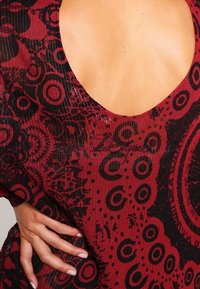 Top a strisce rosse e nere con una profonda scollatura sul retro, caratterizzato da disegni circolari e un tessuto testurizzato. Mano posizionata di lato.